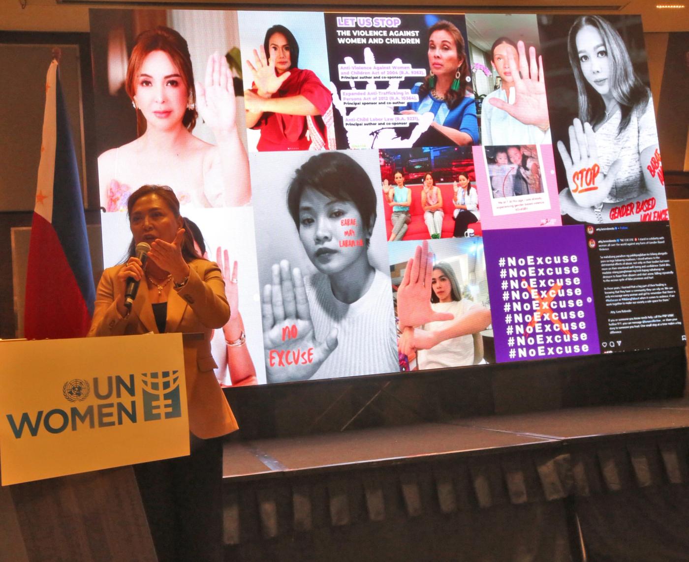 Karen Davila speaks at a UN Women podium before a large screen collage of her #NoExcuse campaign against violence against women and children