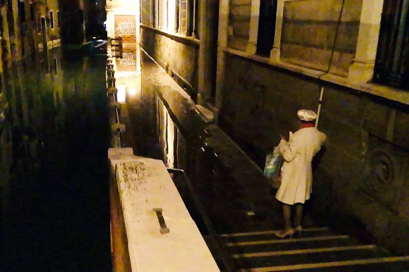 A woman in a white coat on flooded steps beside a Venice canal at night during the acqua alta