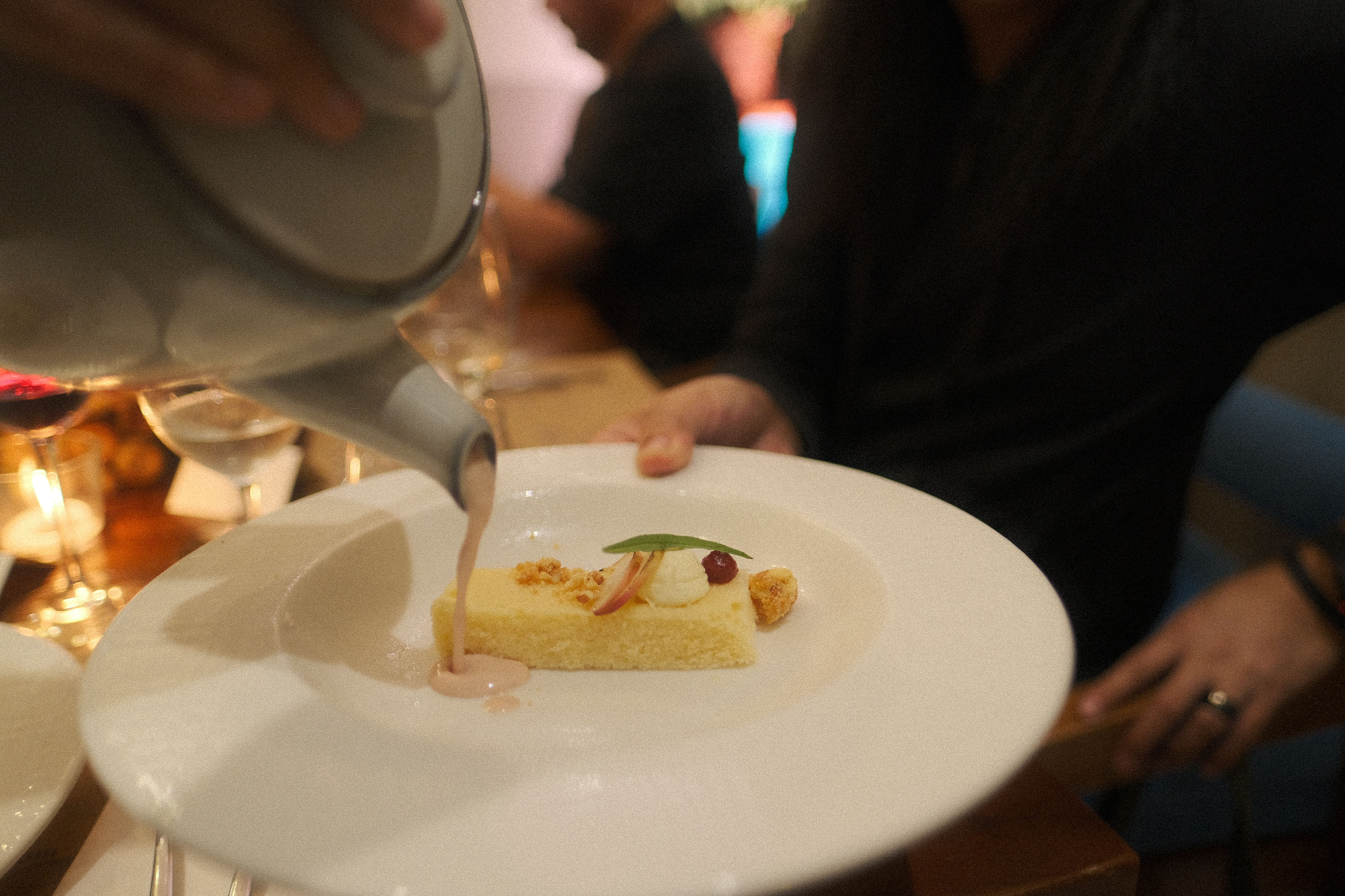 A server pours blush-pink sauce over The Fatted Calf’s peach melba, a sponge slice with cream, fruit and a basil leaf on a white plate