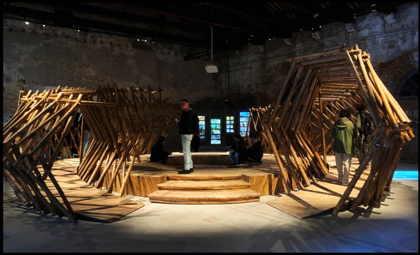 Installation view of ‘Tripa de Gallina: Guts of Estuary’, an angular bamboo lattice tunnel with visitors and a video screen in the brick-walled Arsenale