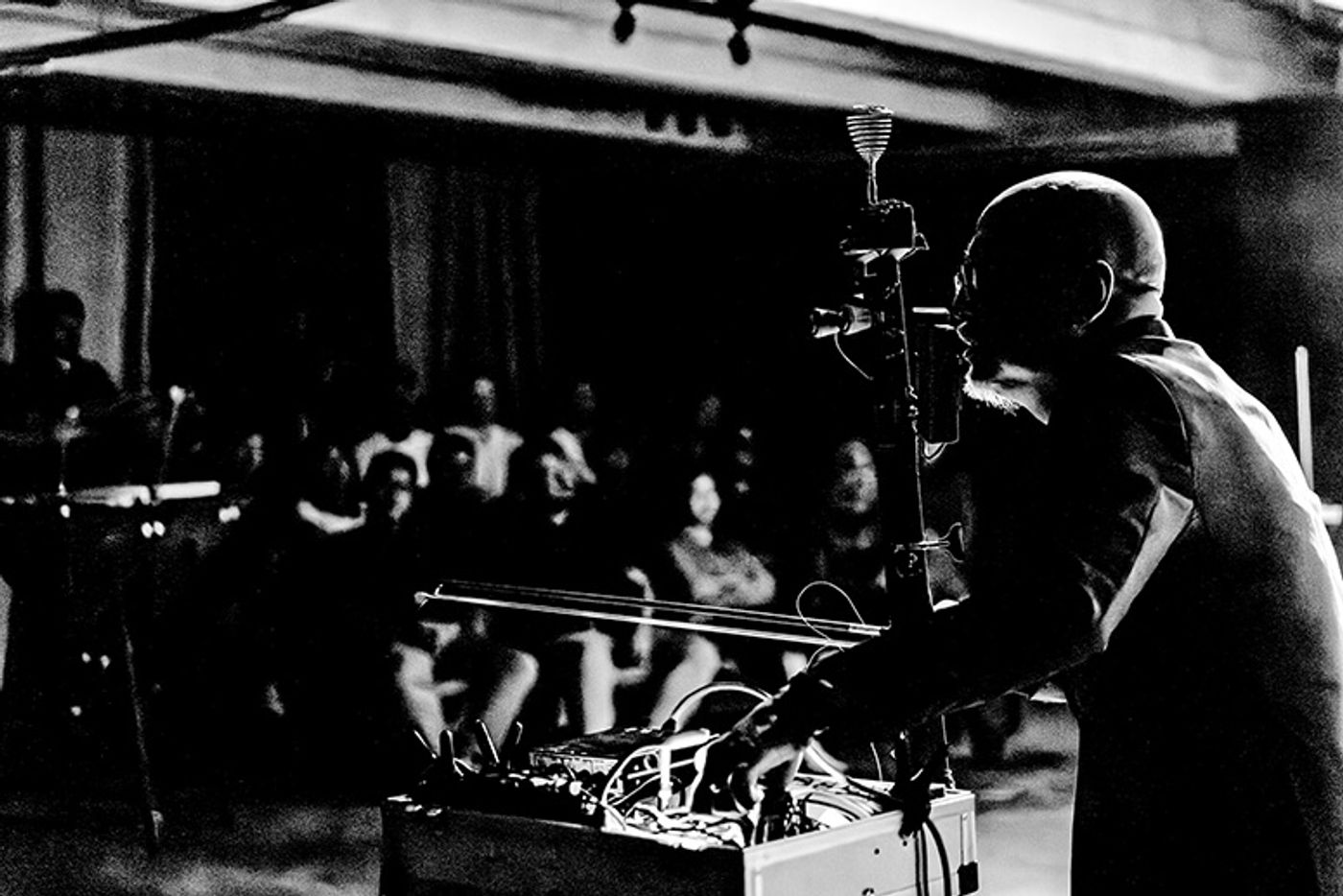 High contrast black and white profile photo of Tad Ermitaño bowing the Tungkod instrument on stage in front of a seated audience
