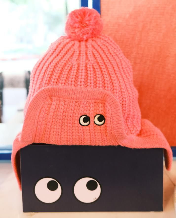 A coral Uniqlo x Anya Hindmarch bobble hat with earflaps and a small pair of embroidered cartoon eyes, resting on a navy box printed with two large cartoon eyes
