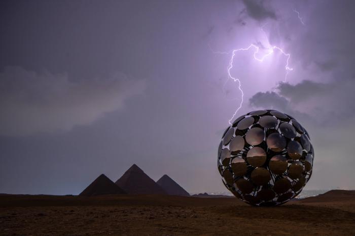 A giant glowing orb of stacked lights rises from the desert plain near the Pyramids of Giza at dusk