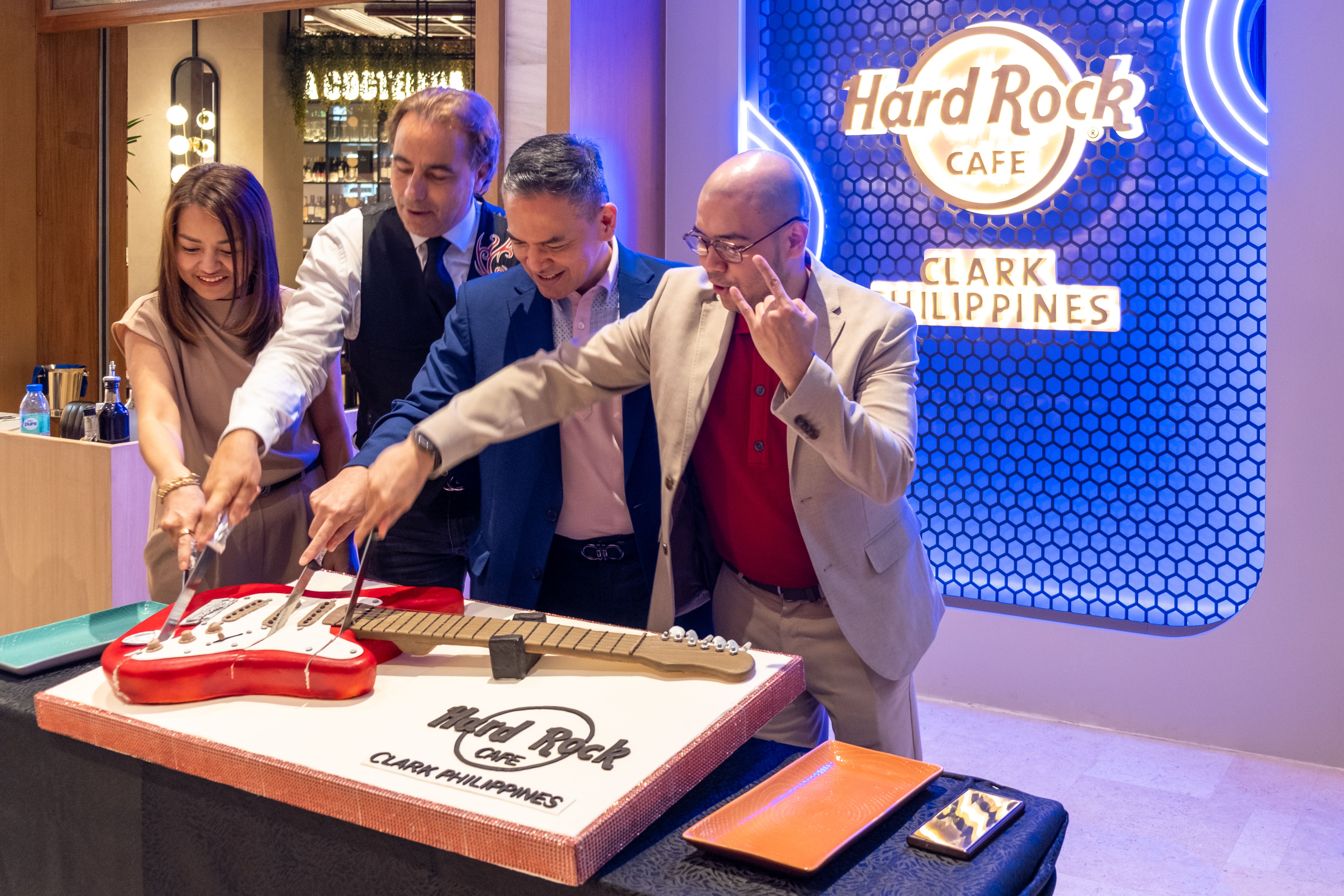 Four people cut into a red guitar-shaped cake beneath the glowing Hard Rock Cafe sign
