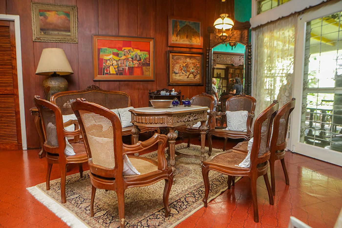 A vintage rattan sala set arranged around a carved centre table on a patterned rug, framed Philippine paintings on the wood-panelled wall above