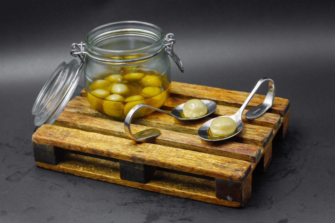 Glass jar of spherified liquid olives on a tiny wooden pallet, two spoons each holding one sphere
