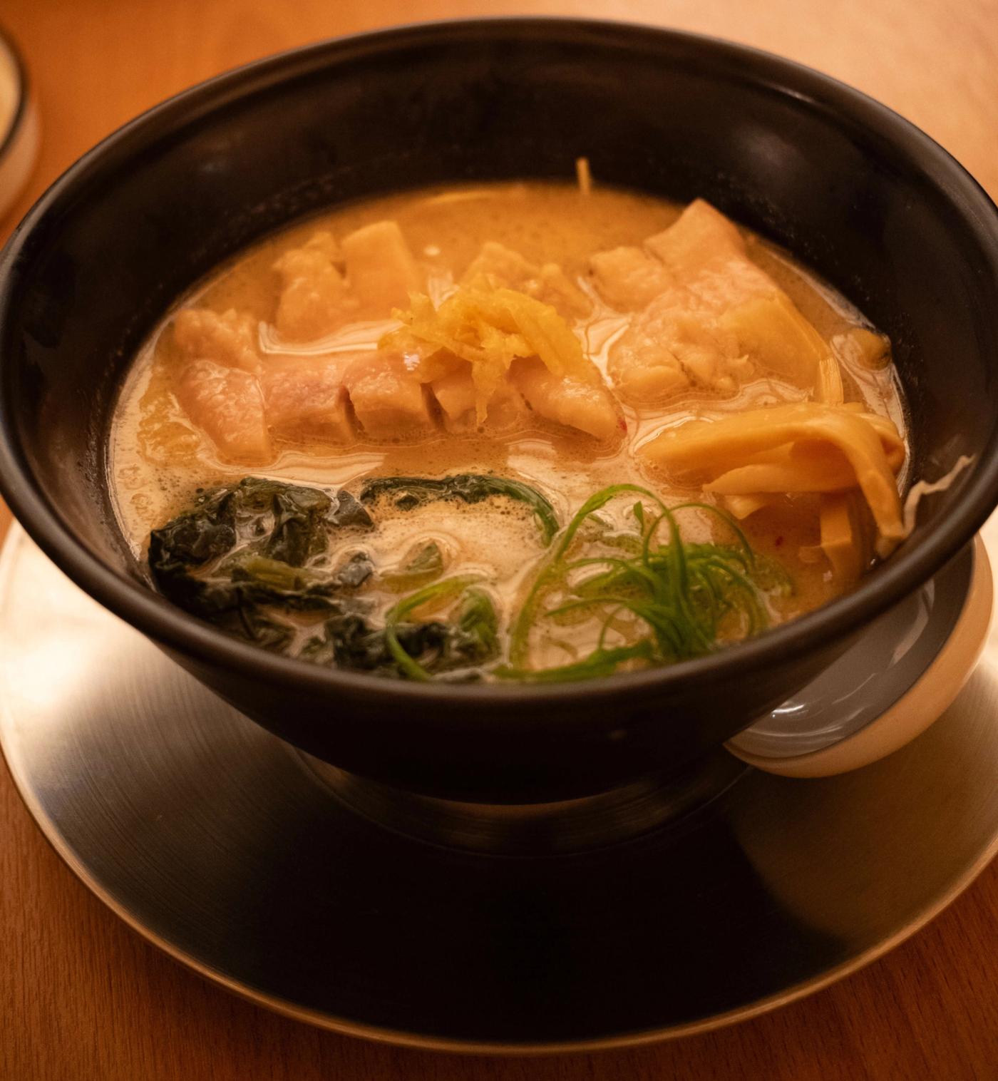 A black bowl of White Chicken Yuzu Ramen: creamy broth with sliced chicken chashu, greens, scallions and bamboo shoots