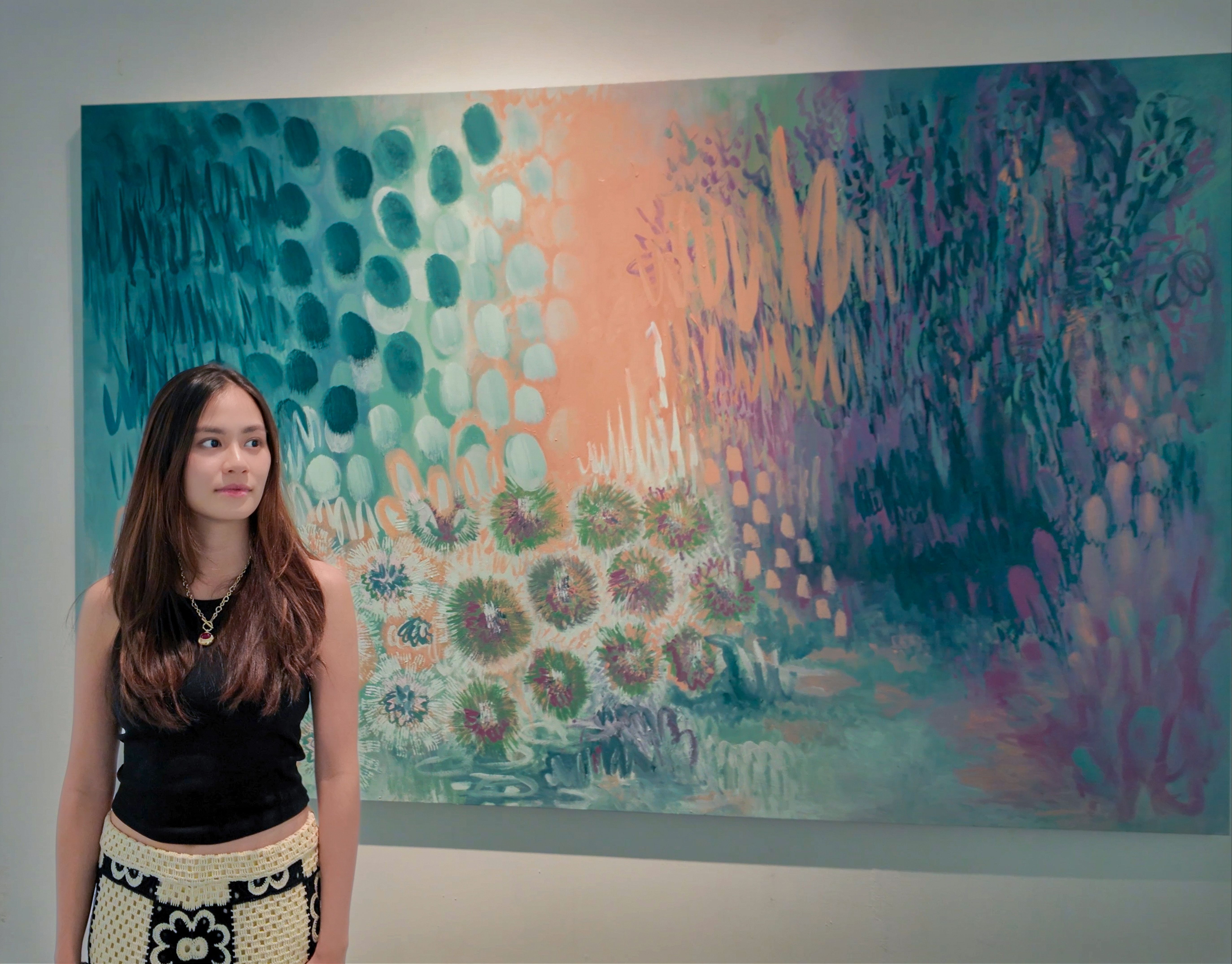 Maria Tan standing beside a large botanical painting layered with teal dots, peach light and clusters of dandelion-like forms