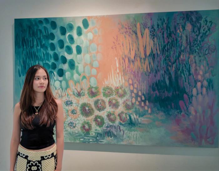 Maria Tan standing beside a large botanical painting layered with teal dots, peach light and clusters of dandelion-like forms