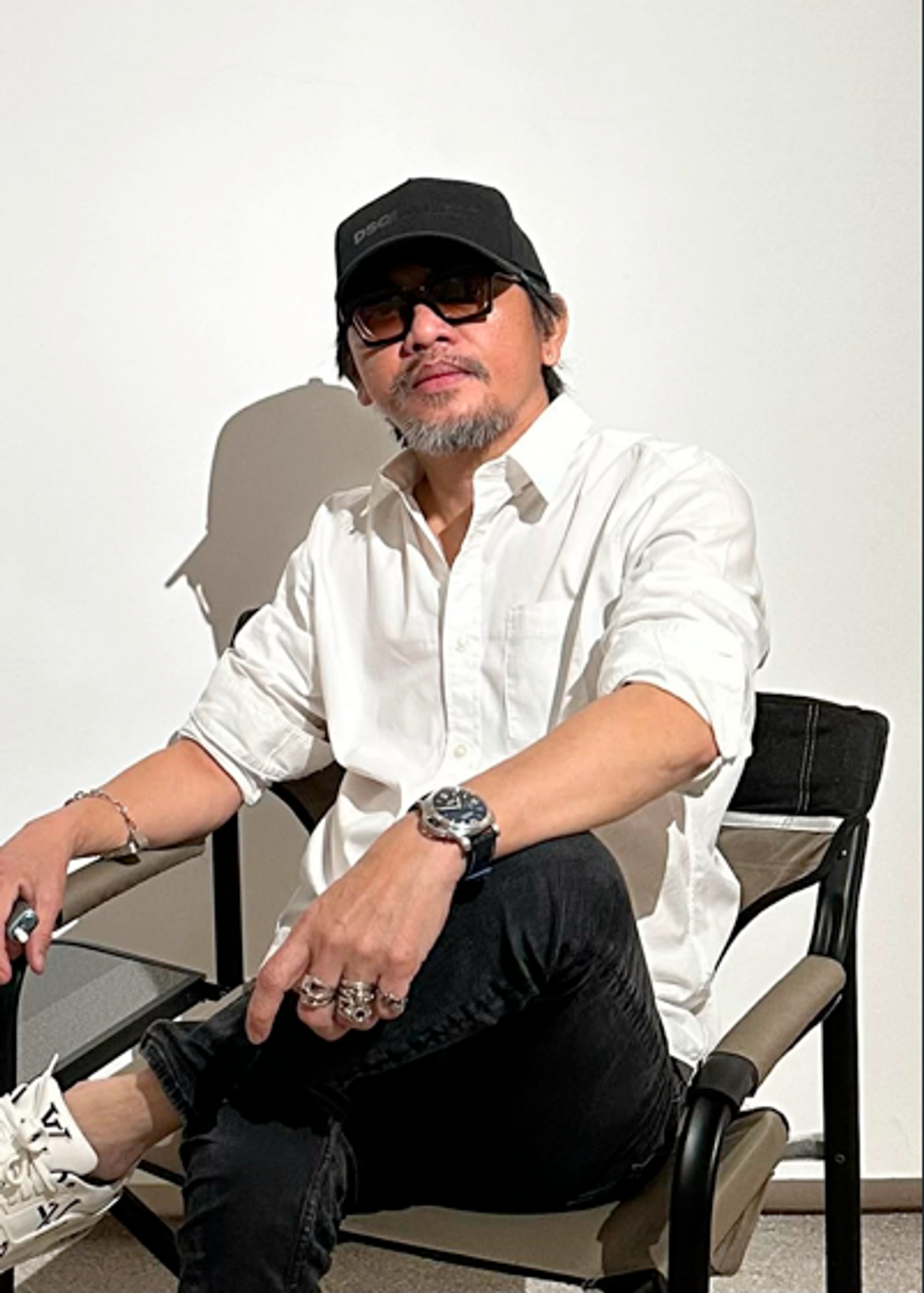 Portrait of Filipino artist Ronald Ventura in a white shirt, black cap and sunglasses, seated with silver rings and a chunky watch