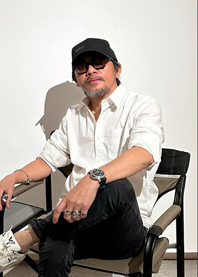 Portrait of Filipino artist Ronald Ventura in a white shirt, black cap and sunglasses, seated with silver rings and a chunky watch