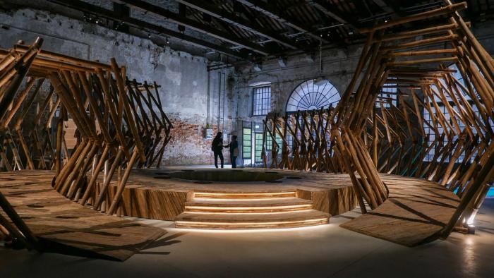 Visitors sit and stand among angular bamboo lattice frames forming a tunnel-like installation in a raw brick-walled hall of the Venice Arsenale