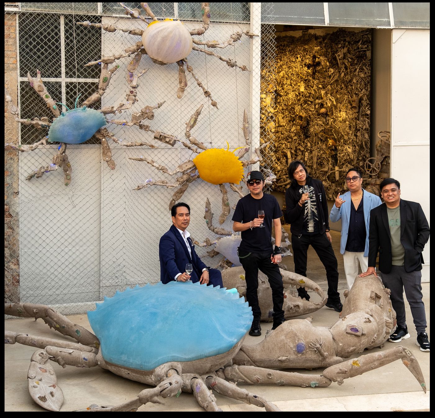 Five men pose among giant crab sculptures with pale blue and yellow shells against a chain-link wall at the Venice docks