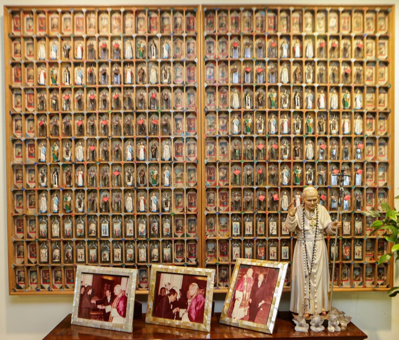 A wall-filling wooden grid of hundreds of small painted saint figures, with framed photographs and a statue of a pope in front