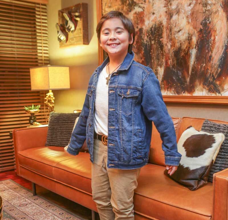 The couple’s young son Diego stands smiling in a denim jacket before a tan leather sofa and a large abstract painting
