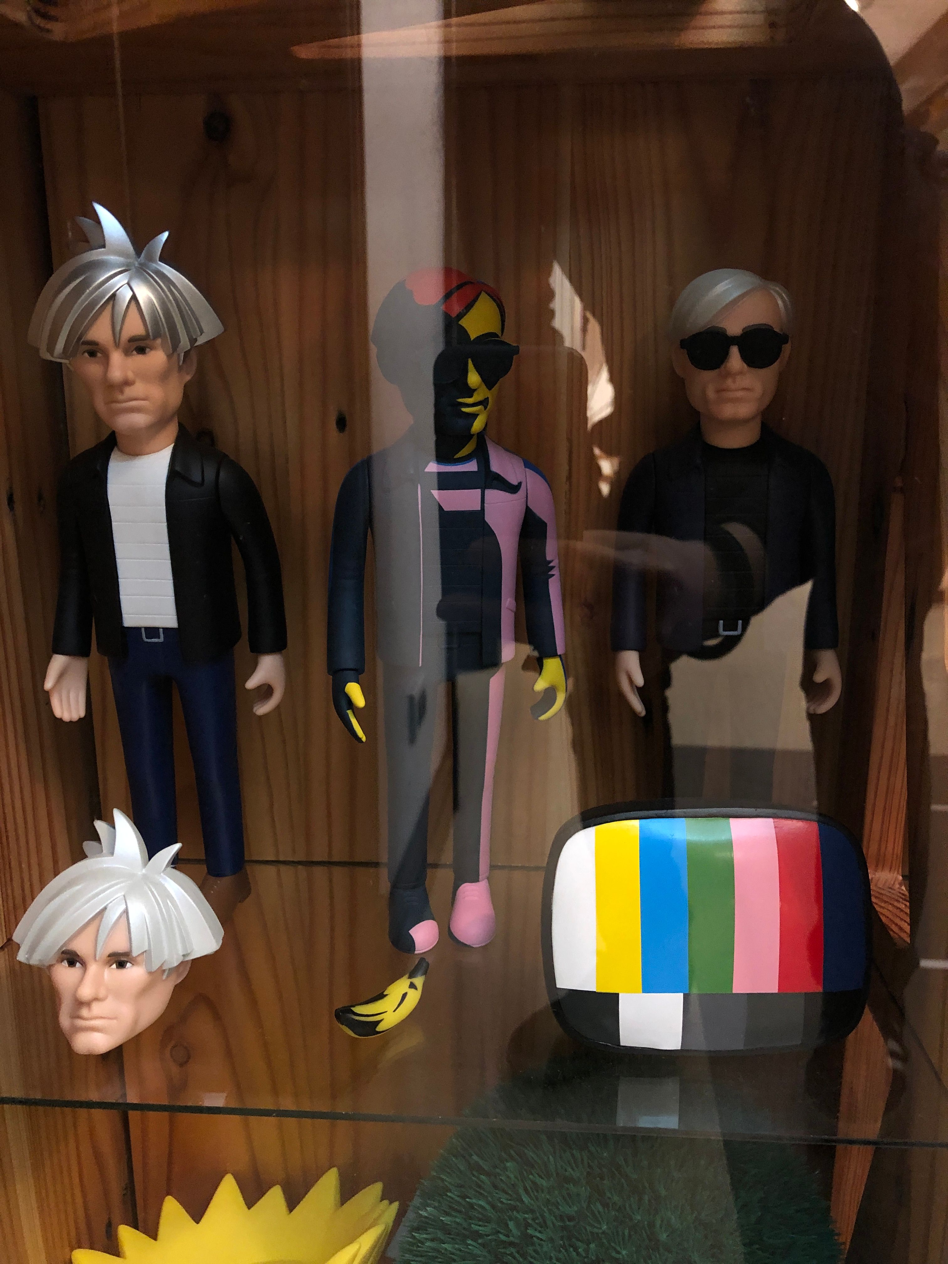 Three Andy Warhol action figures in silver wigs and sunglasses inside a case, with a spare Warhol head, a banana and a colour-bar television toy