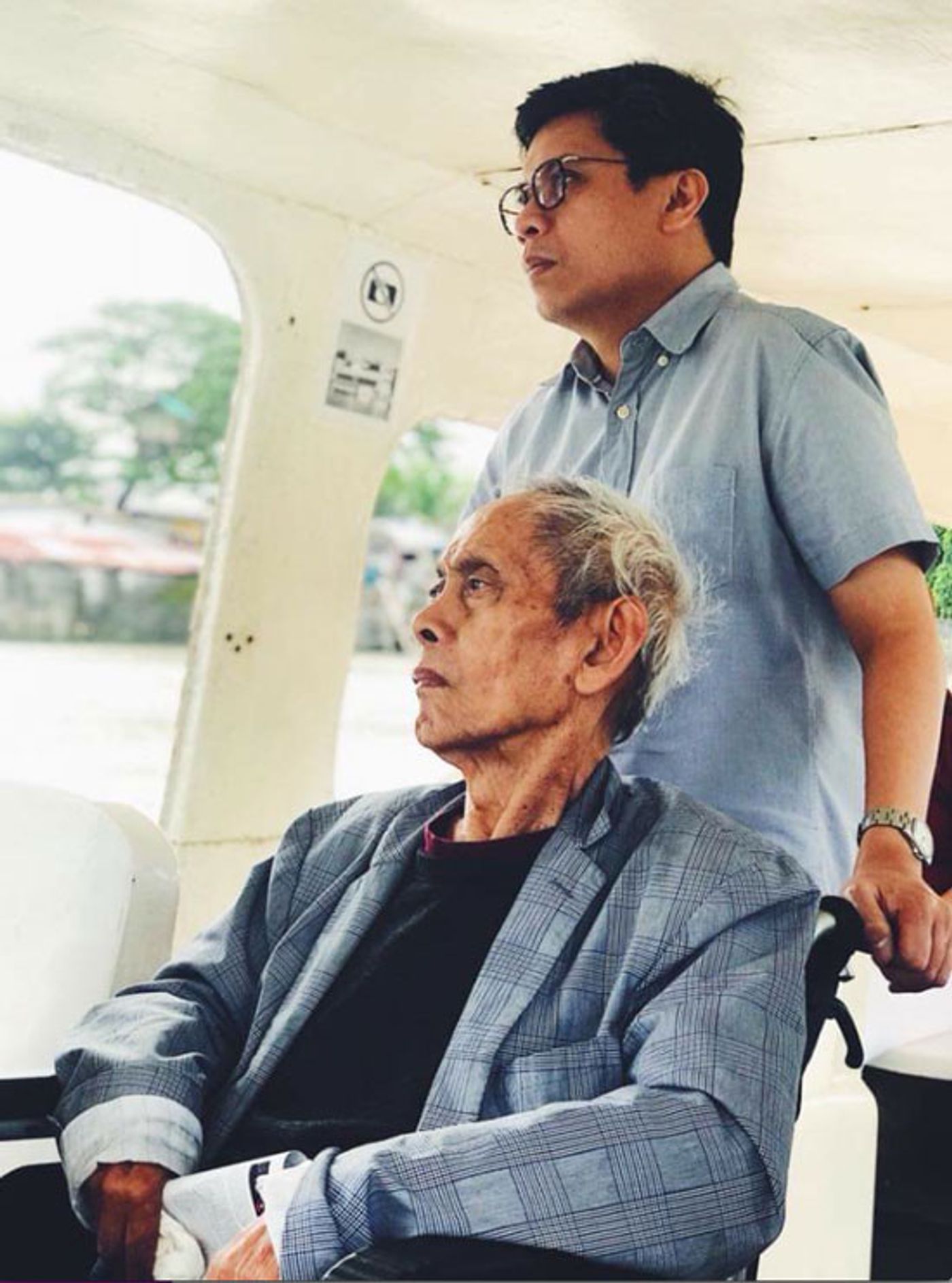 David Medalla seated on a boat with curator Patrick Flores standing behind him