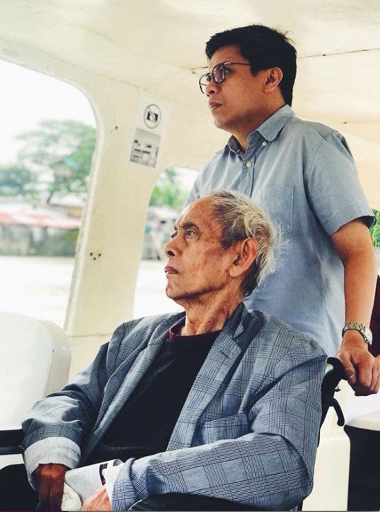 David Medalla seated on a boat with curator Patrick Flores standing behind him