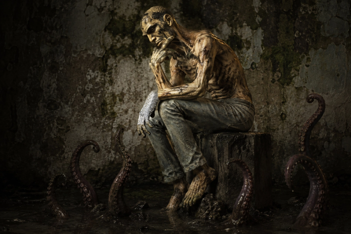 A gaunt, mud-caked man sits in the Thinker pose on a stump, ankle-deep in black floodwater, as octopus tentacles rise around him in a derelict green-stained room