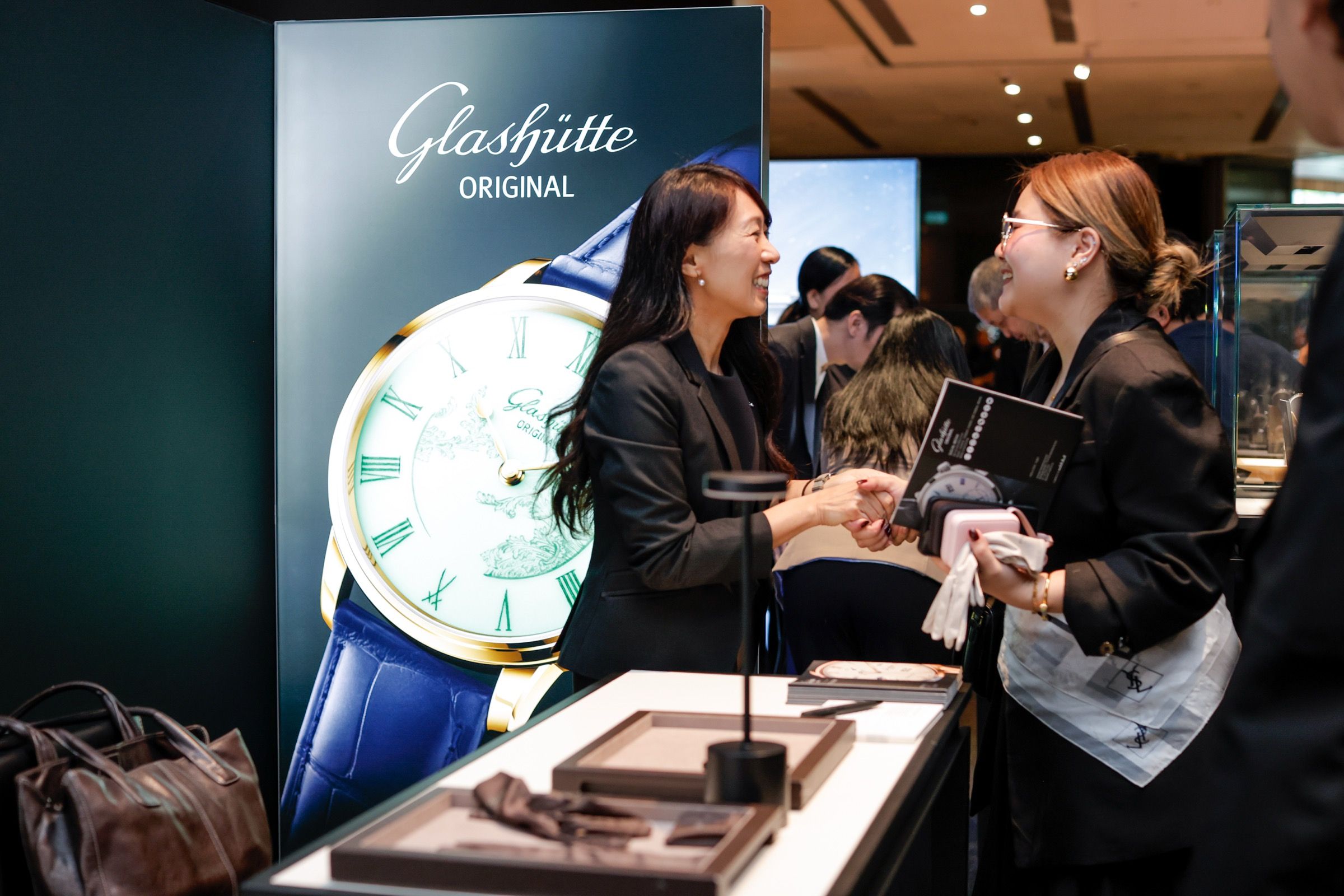 Two women shaking hands smilingly in front of a Glashütte Original watch display backdrop at Lucerne Watch Week
