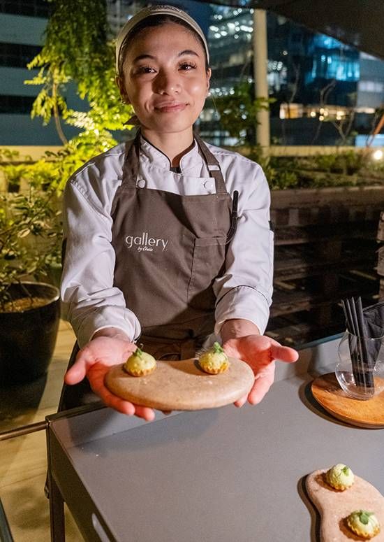 A smiling chef in a Gallery by Chele apron offers two coconut canapes on a stone board at night