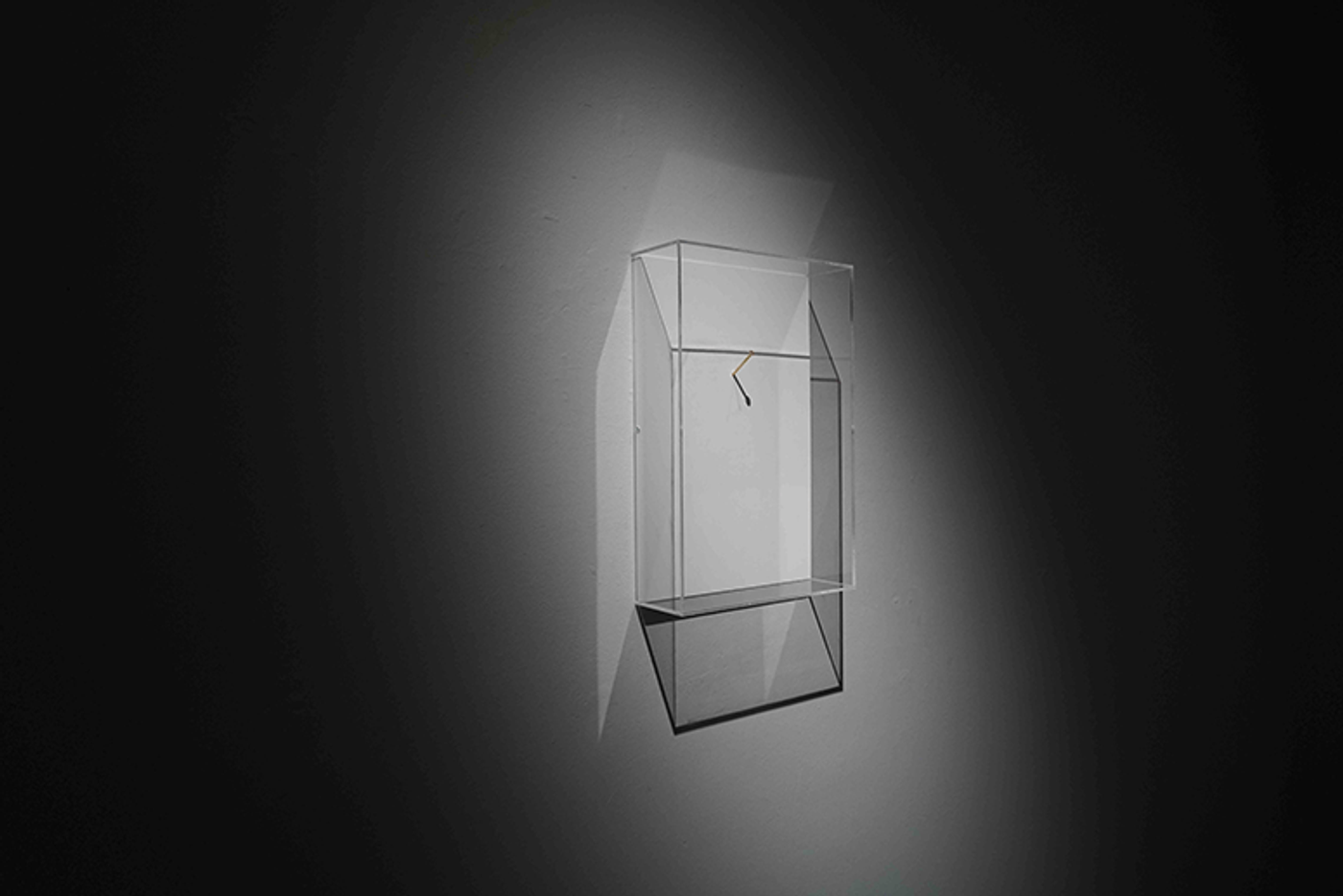 Spotlit clear plexiglass case holding a single gold nail, floating on a dark gallery wall