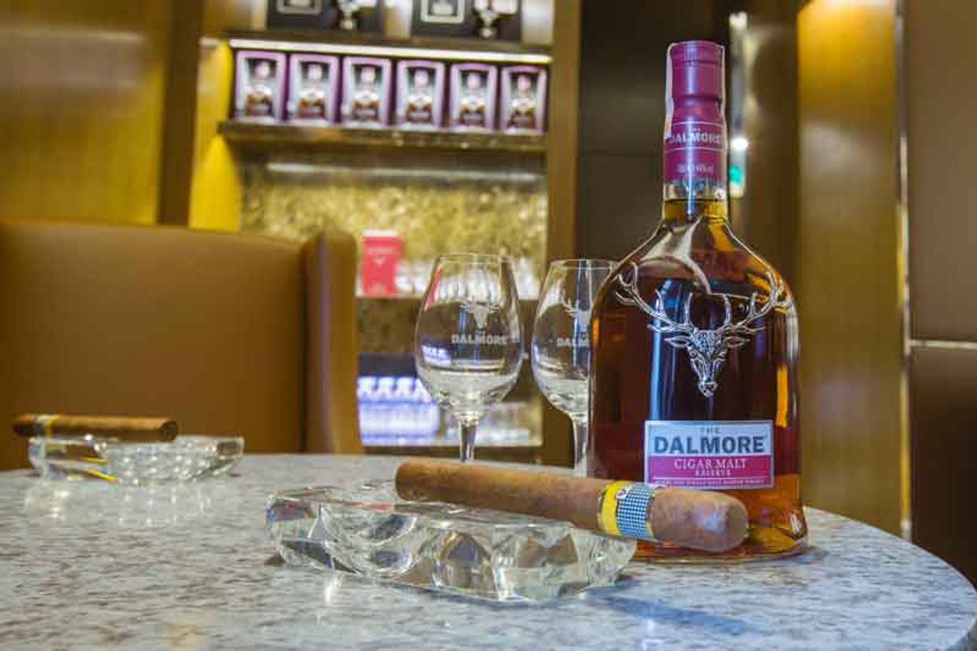A bottle of The Dalmore Cigar Malt Reserve on a marble table with two nosing glasses, a lit cigar and cut-crystal ashtrays