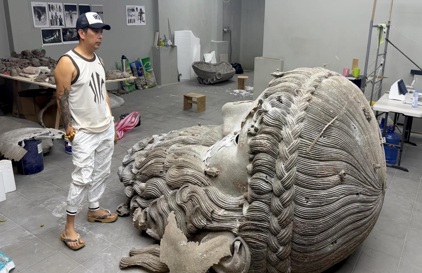 Ronald Ventura beside a monumental fallen head sculpture with thick braided hair in his studio