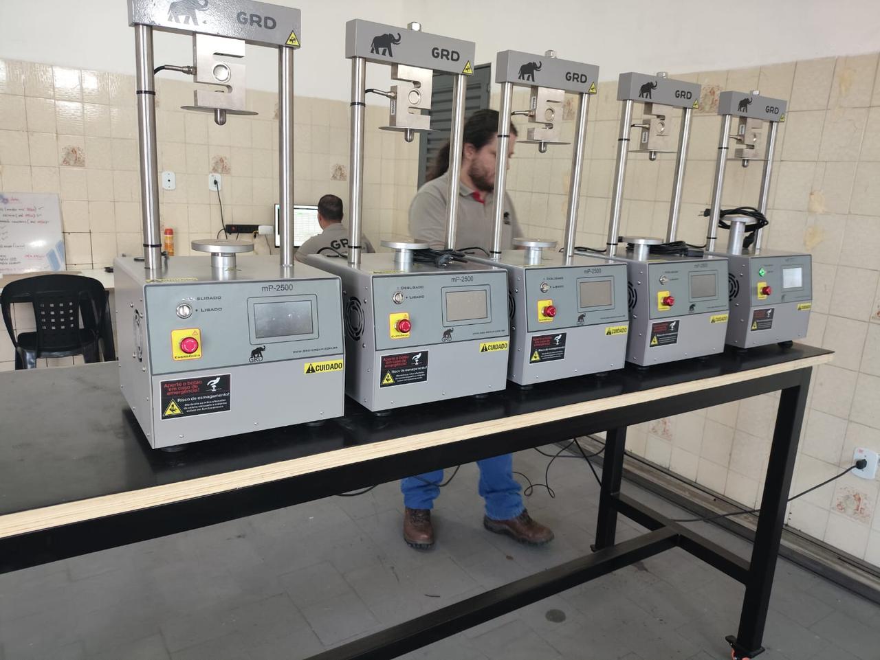 Cinco prensas de bancada GRD alinhadas no laboratório, cada uma com tela e botão de emergência