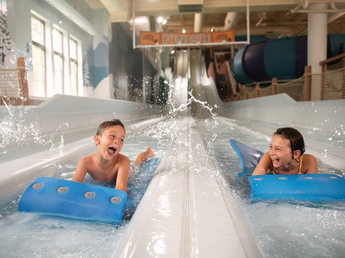 Kids smiling at each other as they race on mats down at waterslide at Speed down a waterslide, float on the Lazy River and relax in a hot tub. You can do it all at Avalanche Bay Waterpark.