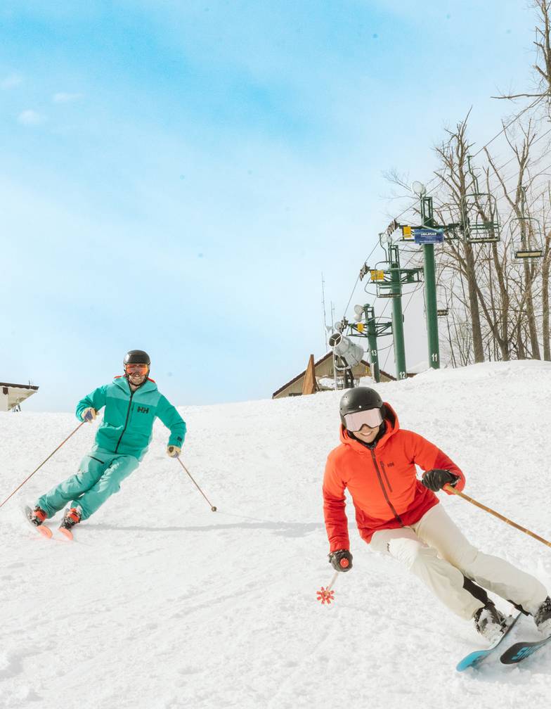 Two skiers in colorful gear carving turns on a sunny groomed run.