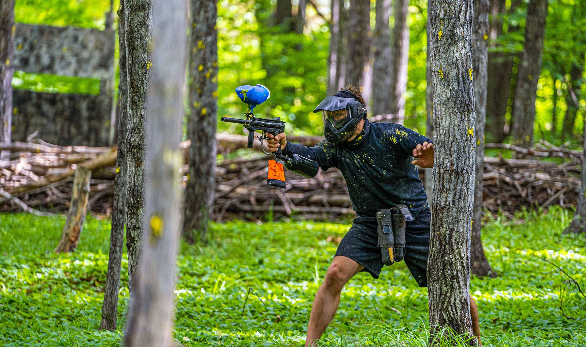 Man hiding behind tree with paintabll gun ready to shoot.