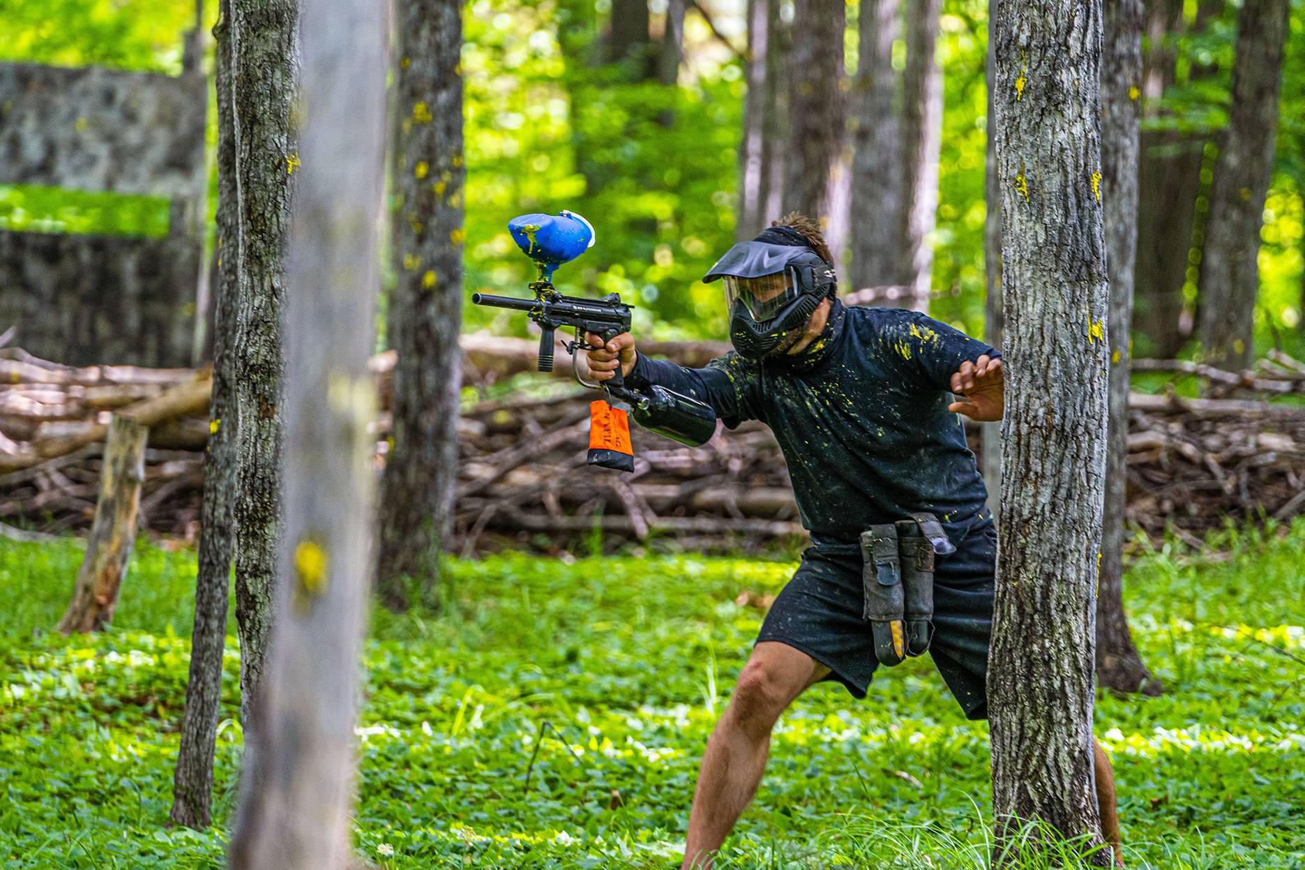 Man hiding behind tree with paintabll gun ready to shoot.