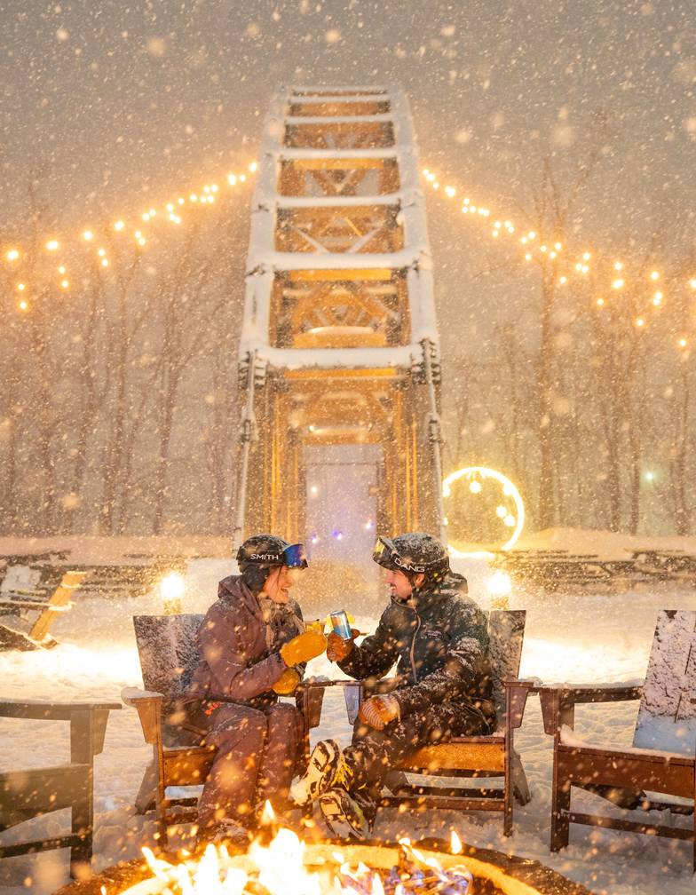 Couple in ski gear clinking drinks by a fire pit in falling snow, with SkyBridge Michigan lit up behind them at night.