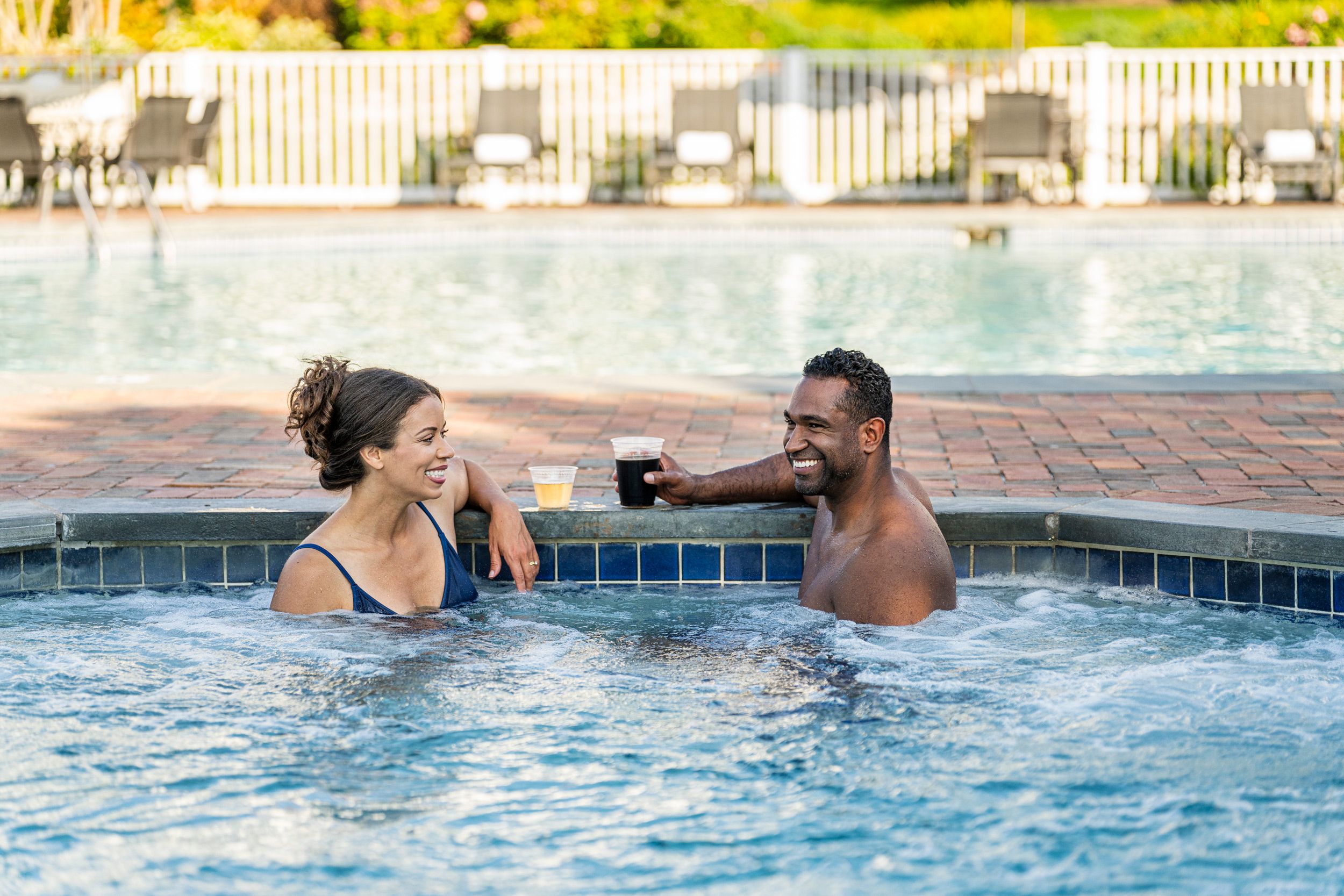 Couple laughing while enjoying drinks in bubbling blue hot tub near outdoor pool