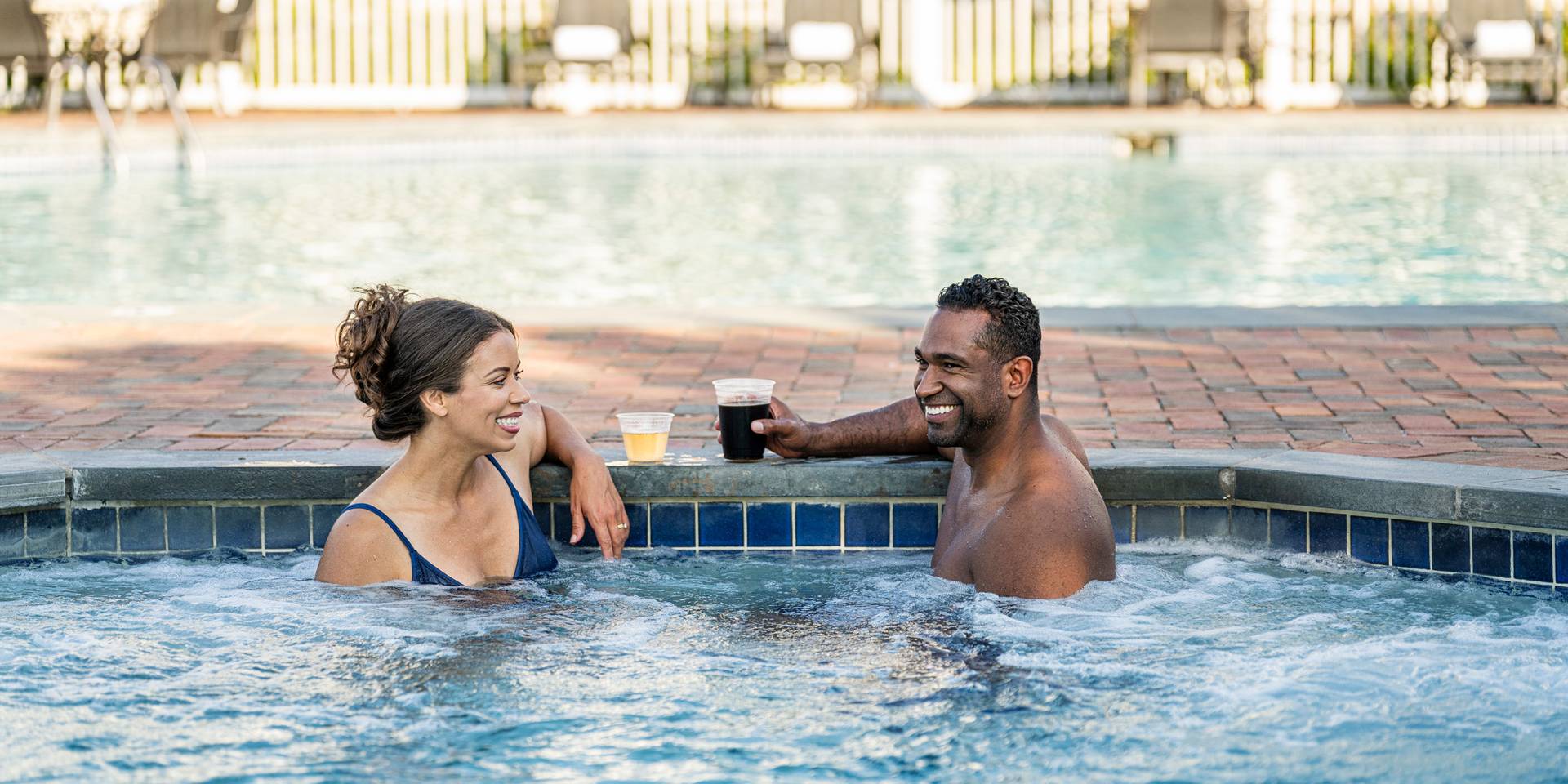 Couple laughing while enjoying drinks in bubbling blue hot tub near outdoor pool