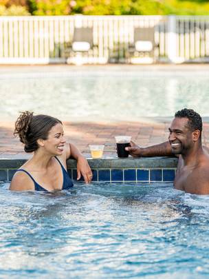 Couple laughing while enjoying drinks in bubbling blue hot tub near outdoor pool