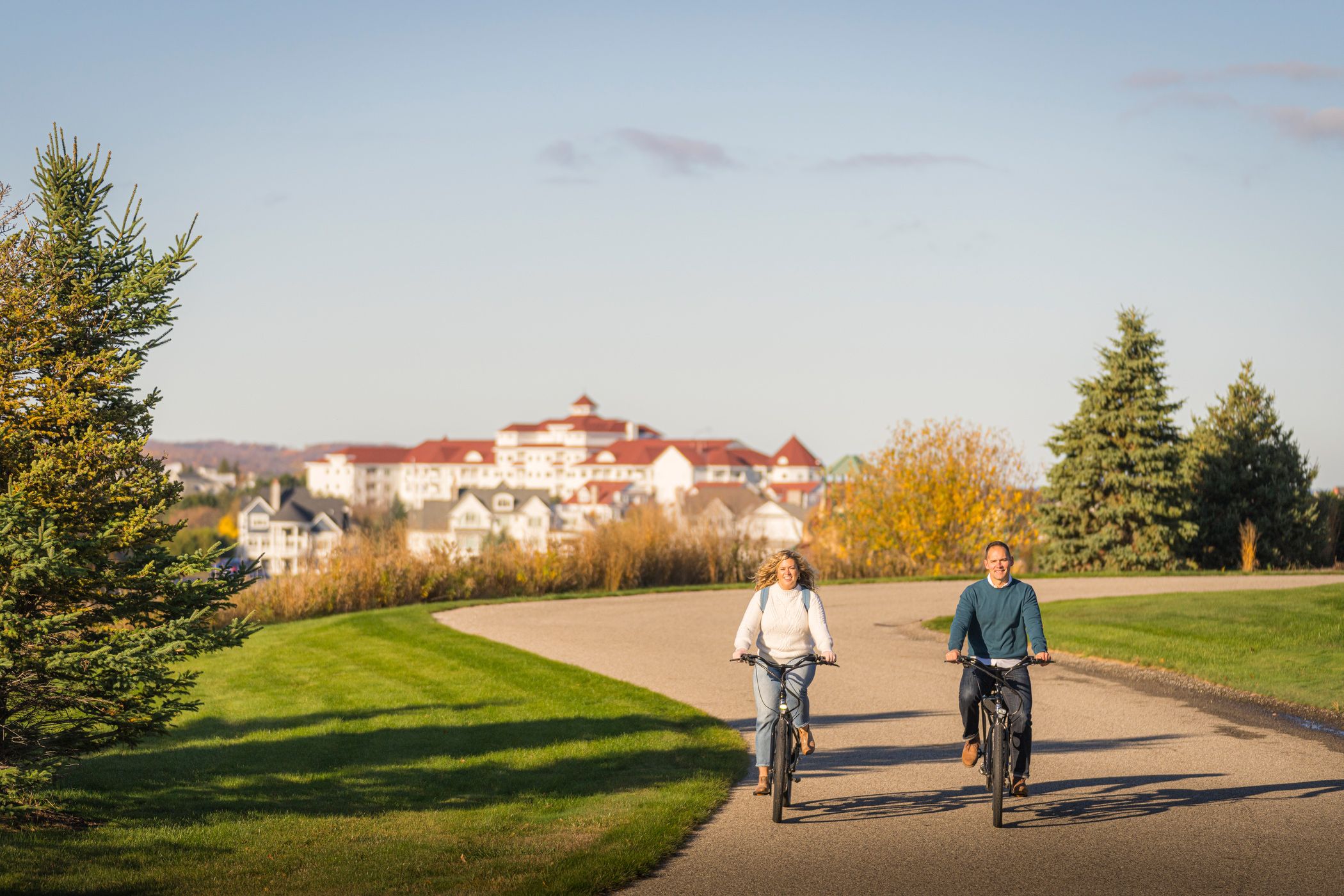 Couple in sweaters rides bikes down winding autumn path with Inn at Bay Harbor in background