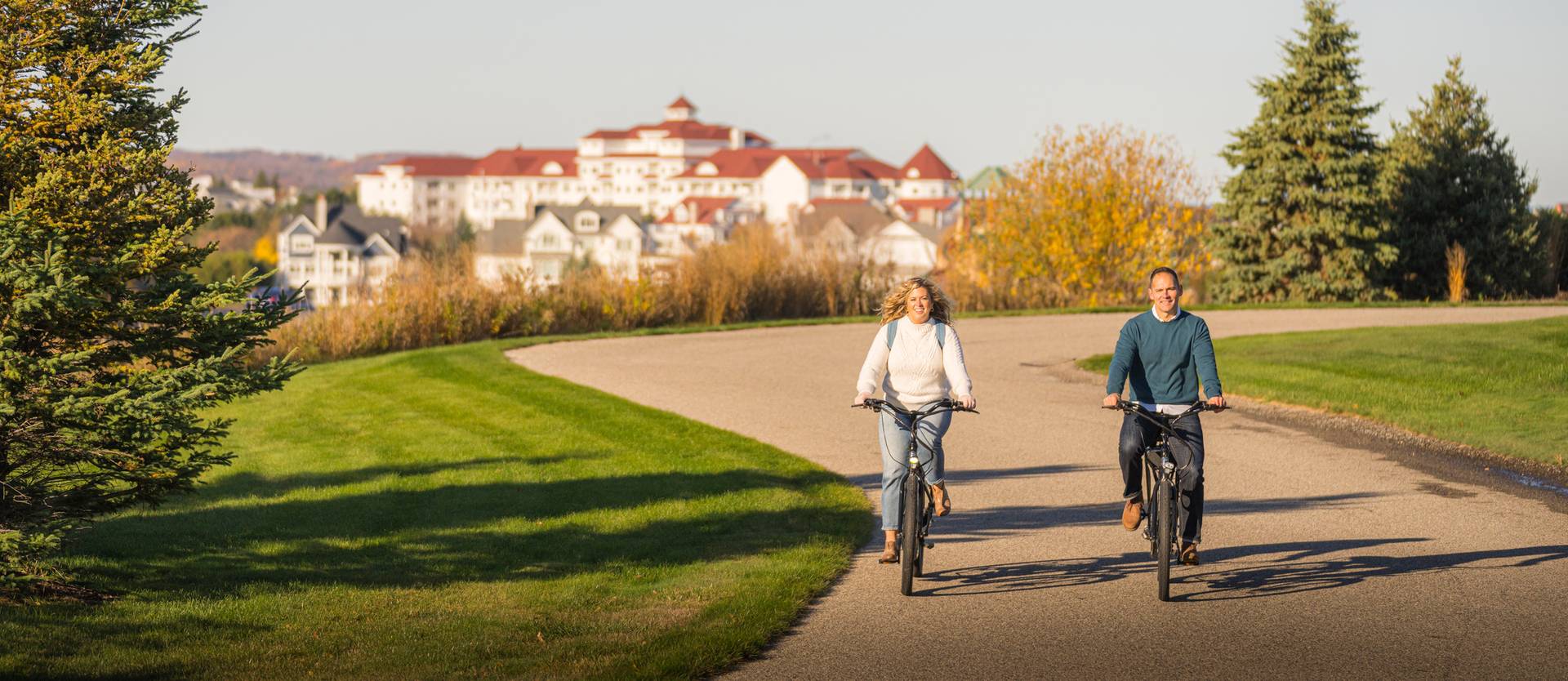 Couple in sweaters rides bikes down winding autumn path with Inn at Bay Harbor in background