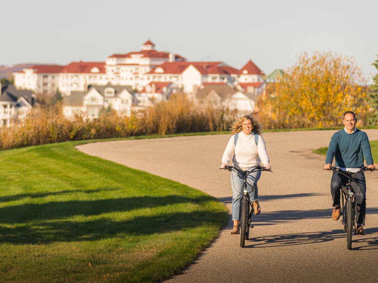 Couple in sweaters rides bikes down winding autumn path with Inn at Bay Harbor in background