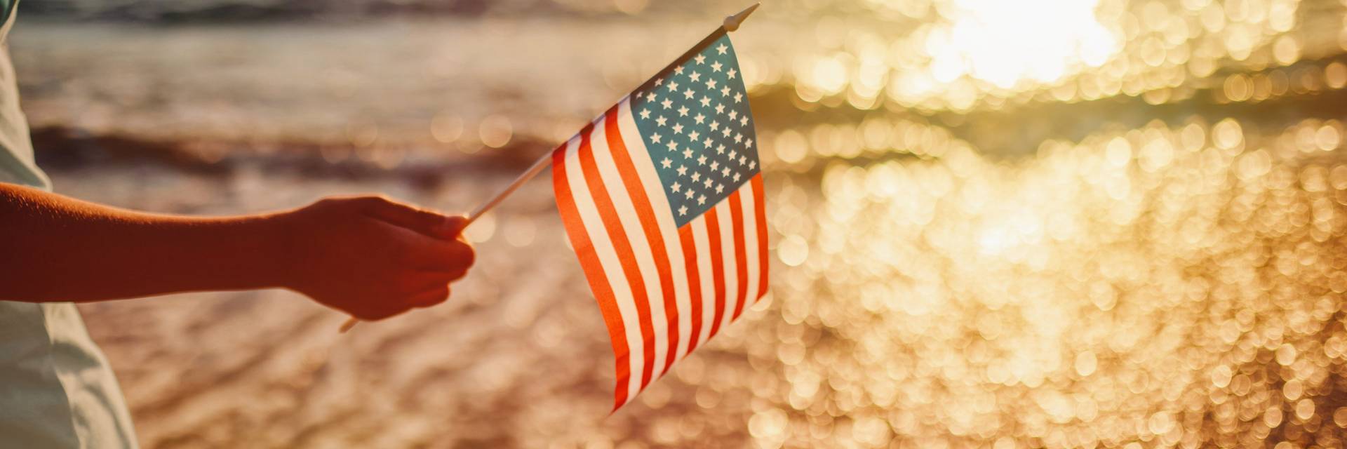 Hand holding American flag near lapping waves of lakeshore at sunset