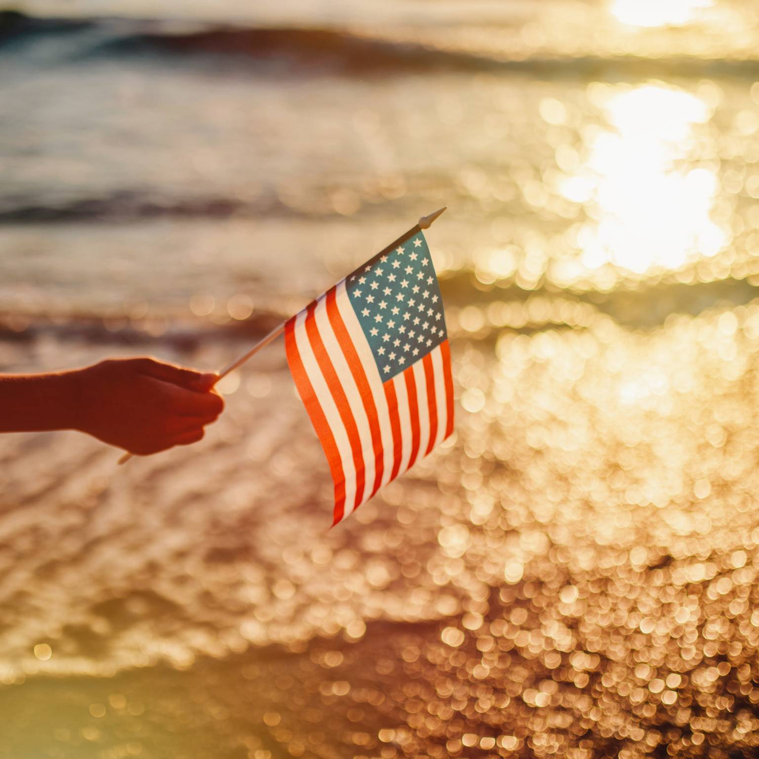 Hand holding American flag near lapping waves of lakeshore at sunset