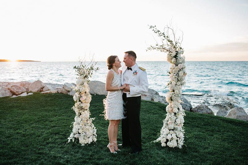 Bride and military groom embrace between floral arches near Lake Michigan at sunset