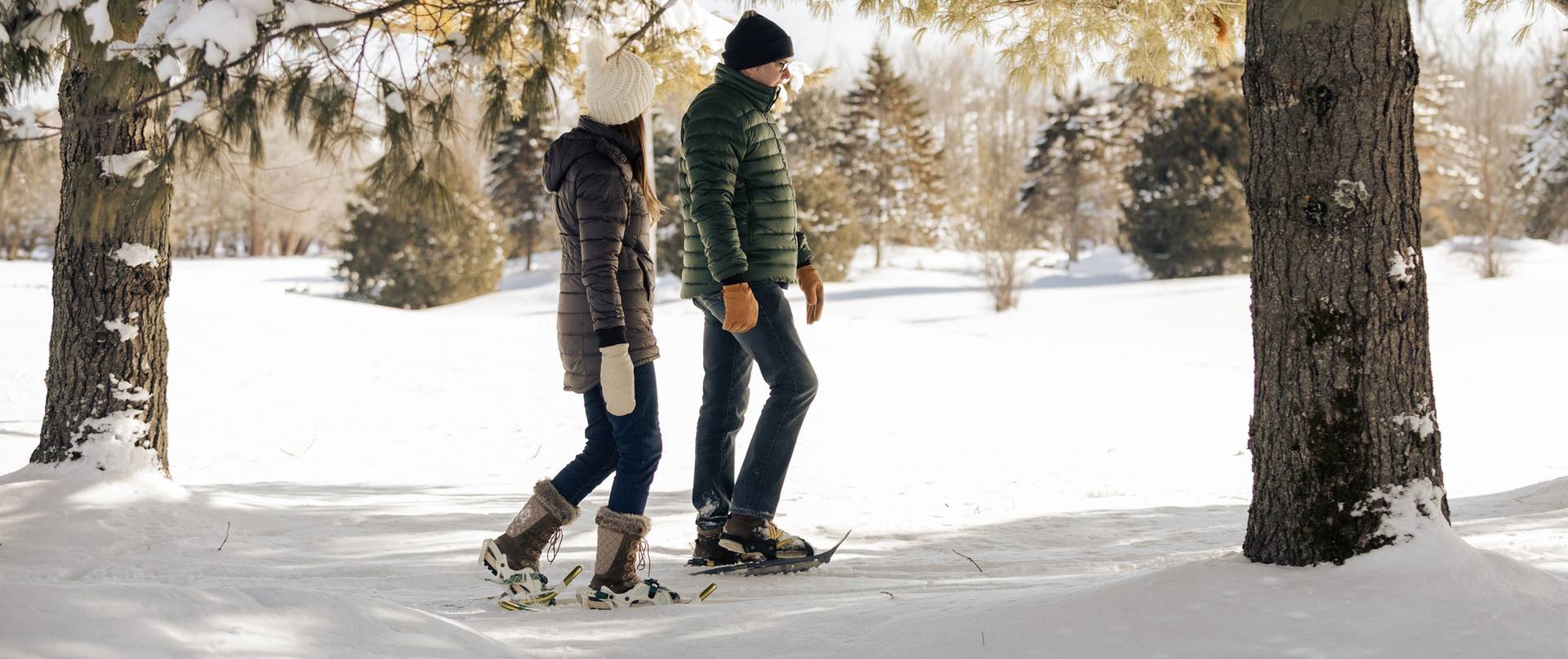 Winter-clad couple walks in snowshoes, chatting, through snowy pine forest