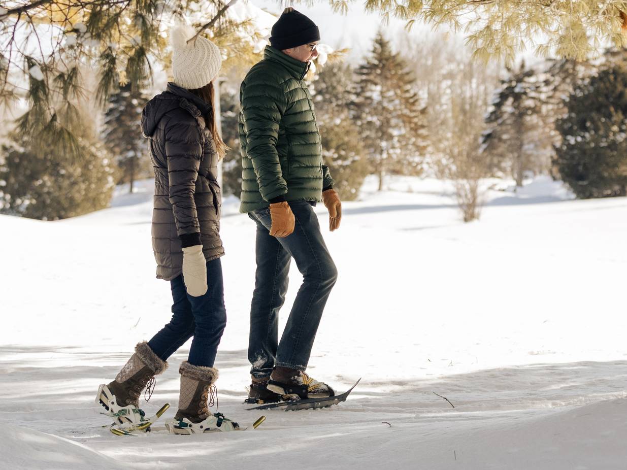 Winter-clad couple walks in snowshoes, chatting, through snowy pine forest