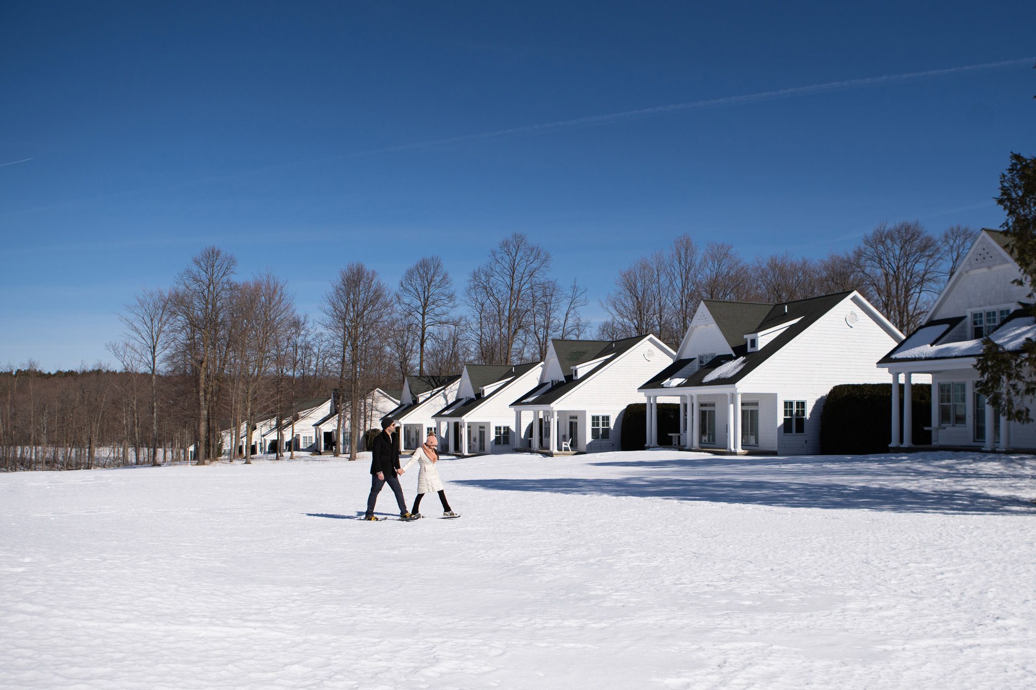 Couple holding hands wearing hats and winter coats treks across snowy landscape lined with Crooked Tree Cottages