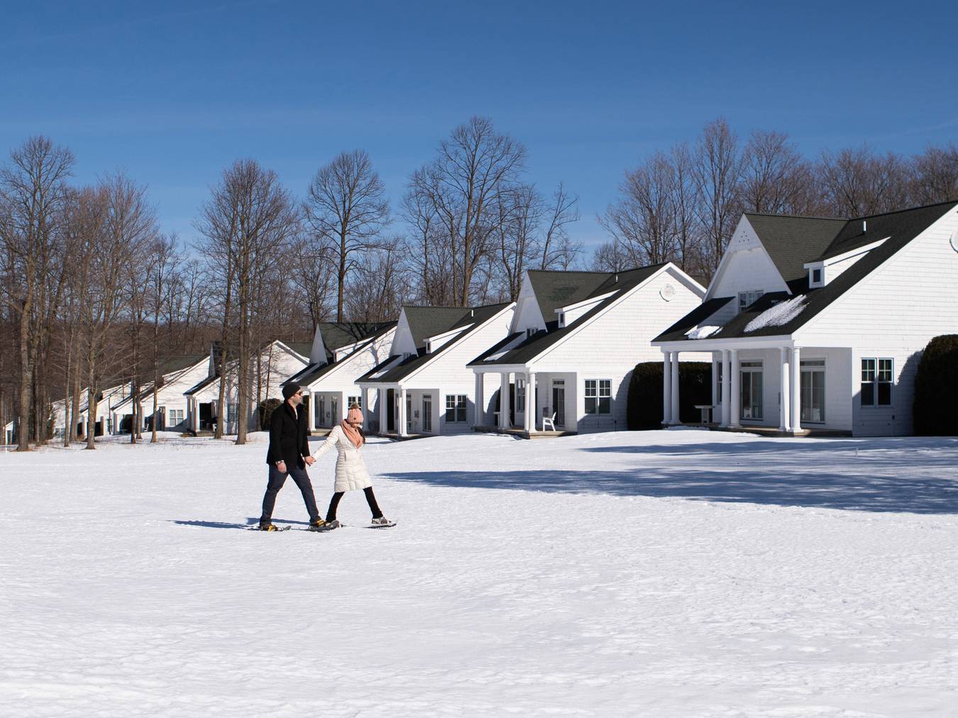 Couple holding hands wearing hats and winter coats treks across snowy landscape lined with Crooked Tree Cottages