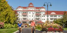 Couple in sunglasses ride bikes down front drive of Inn at Bay Harbor alongside colorful gardens on summer day