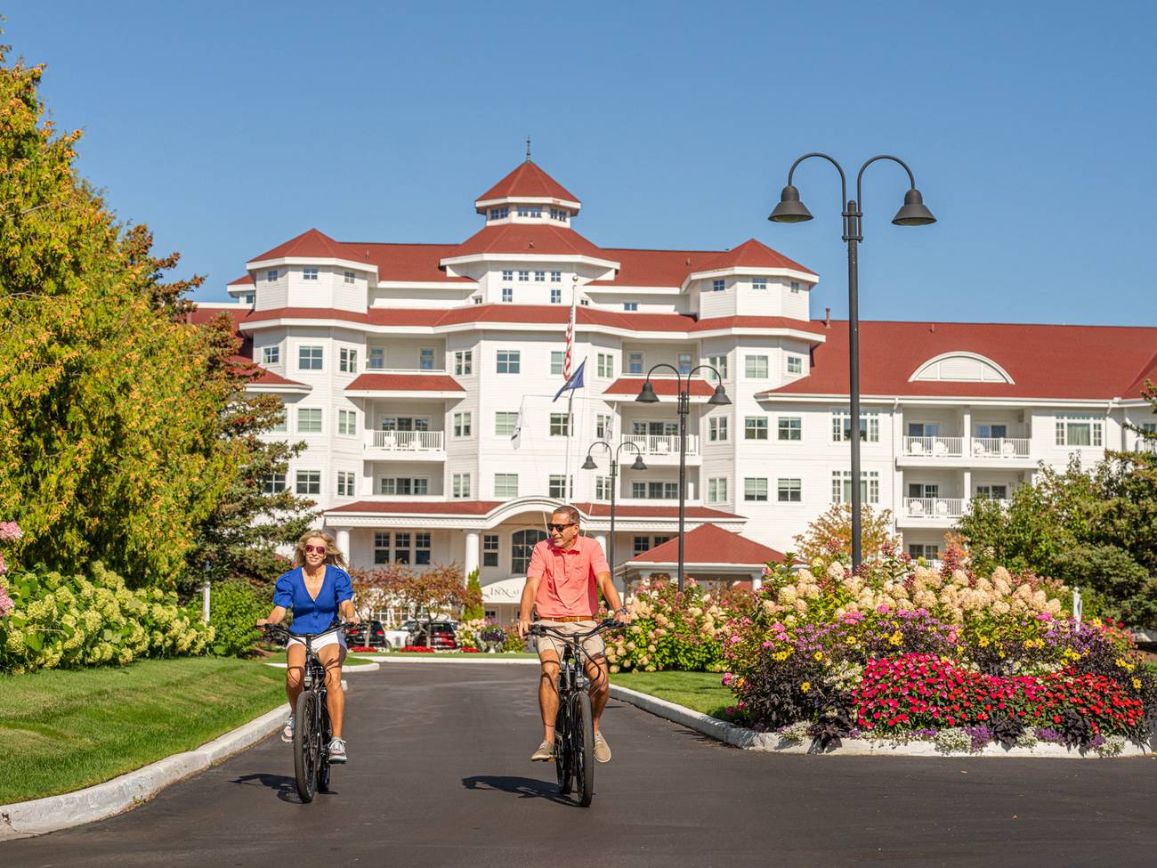 Couple in sunglasses ride bikes down front drive of Inn at Bay Harbor alongside colorful gardens on summer day
