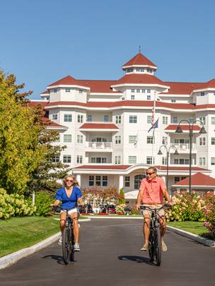 Couple in sunglasses ride bikes down front drive of Inn at Bay Harbor alongside colorful gardens on summer day